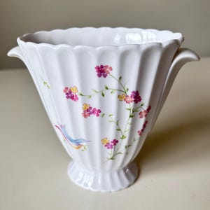 Puede incluir: Jarrón de cerámica blanca con diseño acanalado y detalles florales. El jarrón tiene forma acampanada con borde festoneado y base de pedestal. Delicadas flores rosas, amarillas y moradas y un pájaro azul están pintados en el jarrón.