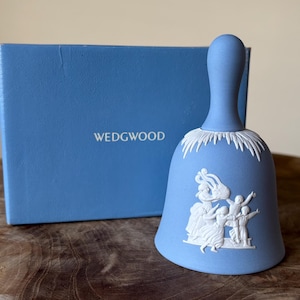 Puede incluir: Campana Wedgwood azul claro con un diseño en relieve blanco de figuras y un ribete blanco decorativo. La campana está junto a una caja azul a juego con la palabra "WEDGWOOD" impresa.