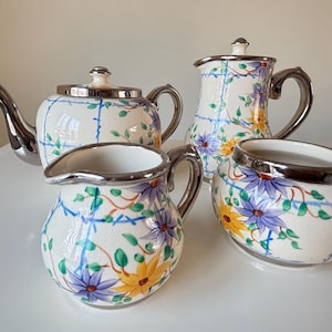 Op de afbeelding: Een vintage theeservies met een theepot, melkkan, suikerpot en een serveerpot. De set heeft een witte basis met een bloemenpatroon in geel en paars, geaccentueerd met groene ranken en blauwe lijnen. Zilveren afwerking.