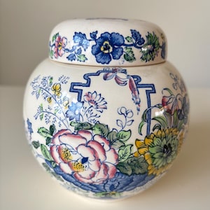 Puede incluir: Un tarro de cerámica redondo con tapa y diseño floral. El tarro tiene una base blanca con patrones florales azules, rosas, amarillos y verdes. La tapa también está decorada con diseños florales azules. El tarro tiene una estética clásica y vintage.