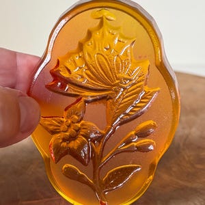 Könnte beinhalten: Ein bernsteinfarbenes, ovales Dekorationselement mit einem Blumen- und Schmetterlingsdesign. Die Reliefschnitzerei zeigt eine detaillierte Blume und einen Schmetterling mit ausgebreiteten Flügeln, vor einem durchscheinenden Hintergrund. Das Stück wird von einer Hand gehalten.