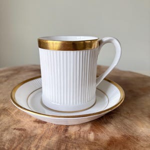 Puede incluir: Un juego de taza y platillo blancos con ribete dorado. La taza tiene detalles acanalados verticales y una banda dorada alrededor del borde. El platillo también tiene un borde dorado y un diseño acanalado a juego. El juego está sobre una superficie de madera.
