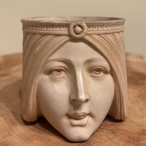 Può includere: Un vaso scolpito color crema a forma di volto femminile. Il volto presenta tratti dettagliati, tra cui occhi, naso e bocca sorridente. La donna indossa una fascia decorativa. Il vaso è probabilmente realizzato in ceramica o materiale simile.