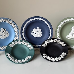 May include: A collection of decorative plates in various colors, including blue, green, and black. Each plate features a white relief design and a white floral border. The plates are displayed on black stands.