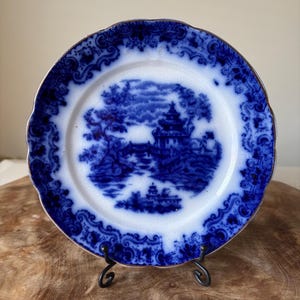 Puede incluir: Un plato antiguo azul y blanco con una escena de paisaje. El plato tiene un borde festoneado con un borde dorado. La imagen central representa una pagoda y árboles. El plato se exhibe en un soporte de metal negro.
