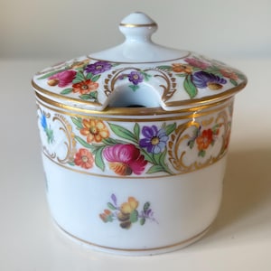 May include: A white porcelain sugar bowl with a lid, adorned with colorful floral patterns and gold trim. The bowl features a small handle on the lid and a notch for a spoon. The floral design includes pink, purple, orange, and blue flowers.