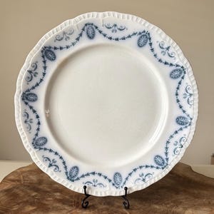 Puede incluir: Plato de cerámica blanca con borde festoneado y diseño floral azul. El plato presenta un patrón ovalado alrededor del borde y se exhibe sobre un soporte de metal negro. Una pieza decorativa de estilo vintage.