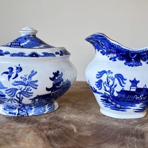 Puede incluir: Un azucarero de cerámica blanco y azul con tapa y una jarra de crema. Ambas piezas presentan un diseño azul de patrón Willow. El azucarero es redondo con tapa, y la jarra de crema tiene asa y pico. El conjunto está sobre una superficie de madera.