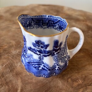 Puede incluir: Una pequeña jarra de crema blanca con un diseño azul de patrón Willow. La jarra tiene un borde festoneado con un ribete dorado y un asa pequeña. El diseño azul representa una escena de paisaje. La jarra probablemente es de porcelana o cerámica.