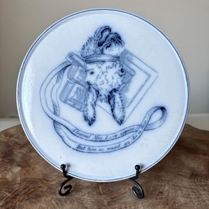 May include: White ceramic plate with a blue illustration of a sheep wearing a crown, with books and a banner that reads "A learned Man I now appear, But turn me round an Ass is there." The plate is displayed on a black metal stand.