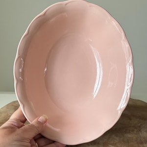 May include: A pale pink ceramic bowl with a wavy edge. The bowl is circular and has a smooth, shiny surface. The bowl is held by a hand, and the background is a light green colour.
