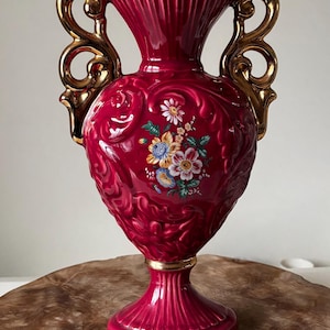 Puede incluir: Un jarrón de cerámica burdeos con detalles dorados. El jarrón presenta asas ornamentadas y un diseño floral. La base y el borde están adornados en oro. El jarrón mide aproximadamente 30 cm de alto.