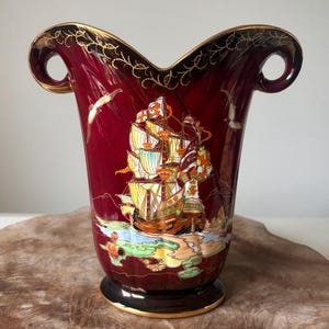May include: A maroon vase with a gold rim and handles. The vase features a detailed illustration of a sailing ship with colourful sails and a coastal scene. Decorative gold accents and flying birds enhance the design.