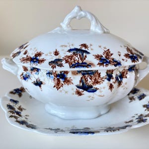 Op de afbeelding: Een witte porseleinen juskom met een deksel en een bijpassende schotel. De juskom en de schotel zijn versierd met een bloemmotief in tinten blauw en bruin.