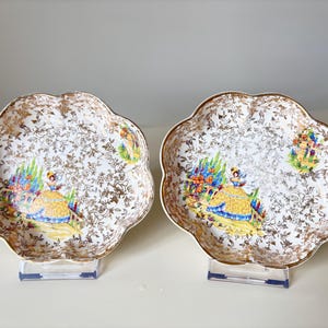 Puede incluir: Dos platos decorativos vintage con bordes festoneados. Las superficies blancas están cubiertas con un patrón floral dorado. Cada plato presenta una ilustración colorida de una mujer con un vestido amarillo y una escena de jardín. Los platos se exhiben sobre soportes transparentes.