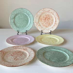 Puede incluir: Seis platos de cerámica vintage en colores pastel: verde, melocotón, rosa, amarillo y dos tonos de verde. Cada plato tiene un borde dorado y un patrón texturizado único. Los platos se exhiben sobre una superficie blanca.