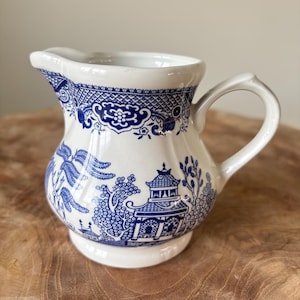 May include: A white and blue ceramic creamer with a classic Willow pattern. The creamer features a spout, a curved handle, and a detailed blue design of a pagoda, trees, and other elements. The creamer is approximately 4 inches tall.