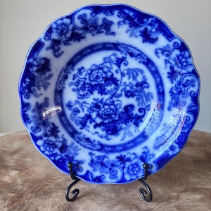 May include: A blue and white antique plate with a floral design. The plate has a scalloped edge and is displayed on a black metal stand. The intricate pattern features flowers and foliage in shades of blue against a white background.