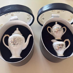 May include: Two black boxes with lids, each containing a miniature tea set. The tea sets are white with gold accents and floral designs. The boxes are labelled "Coalport" and "England".