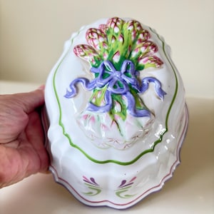 May include: White ceramic mould with a raised design of a bouquet of asparagus tied with a purple ribbon. The mould has a scalloped edge and green, purple, and white accents.