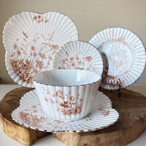 Puede incluir: Un juego de vajilla de porcelana blanca con un estampado floral en tonos marrones. El juego incluye platos y un cuenco, todos con borde ondulado y ribete dorado. La vajilla está dispuesta sobre una superficie de madera, mostrando una estética vintage.