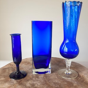 Può includere: Tre vasi di vetro blu cobalto di varie forme e dimensioni. Il vaso a sinistra è un piccolo vaso a bocciolo. Il vaso centrale è rettangolare. Il vaso a destra ha una base bulbosa e un bordo increspato.