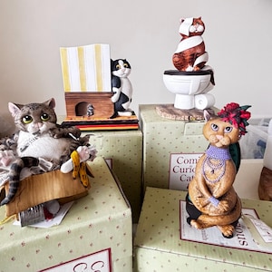May include: A selection of quirky cat ornaments. One cat is seated, holding a newspaper and a banana peel. Another cat is perched on a toilet. A third cat peeks from behind a structure, and the last cat is adorned with a floral headpiece and jewellery.