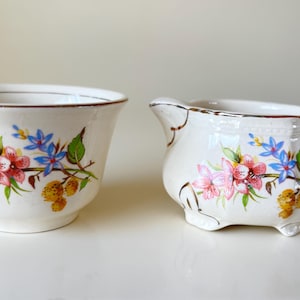 Könnte beinhalten: Ein weißes Keramik-Set aus Milchkännchen und Zuckerdose mit Blumenmuster. Das Milchkännchen hat einen Henkel und einen abgerundeten Körper. Die Zuckerdose ist rund und hat einen goldenen Rand.