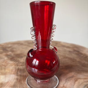 May include: A deep red glass vase with a rounded base and a flared top. The vase features clear glass handles and rests on a clear, circular base. The vase is approximately 18 cm tall.