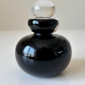 May include: A small, round, black glass perfume bottle with a clear, spherical stopper. The bottle has a glossy finish, reflecting light. The design is simple and elegant, suitable for storing fragrances or essential oils.