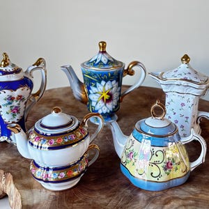 Puede incluir: Cinco teteras decorativas con intrincados diseños florales y detalles dorados. Las teteras varían en forma y color, incluyendo azul, blanco y amarillo, y están dispuestas sobre una superficie de madera. Cada tetera tiene una tapa y un asa.