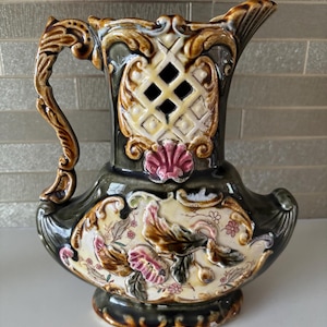 May include: A green and brown ceramic jug with intricate floral and scrollwork designs. The jug has a lattice-like opening on the side and a decorative handle.