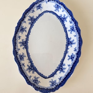 May include: An oval ceramic platter featuring a white centre and a wide, scalloped border decorated with a blue floral pattern and a geometric Greek key design. The surface displays a fine network of crazing throughout the white glazed area.