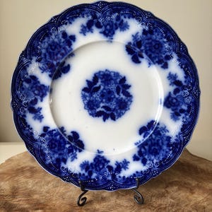 May include: A white and blue floral patterned plate with a scalloped edge. The plate features a central floral design and a border of blue floral patterns. The plate is displayed on a black metal stand, set against a wooden surface.