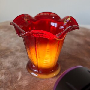 May include: A red and orange glass vase with a ruffled rim. The vase has a flared shape and a round base. The text "OSBURN ABBEY" is visible on the rim, along with small floral accents. The vase is likely a decorative item.