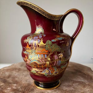 May include: A maroon ceramic jug with a gold rim and handle. The jug features a detailed landscape scene with colourful buildings and trees. The base is black with a gold trim.