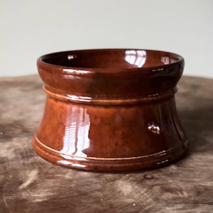 Può includere: Un contenitore in ceramica marrone con una base ampia e svasata e una parte superiore più stretta. Il contenitore ha una finitura lucida e una fascia di colore più chiaro al centro. L'interno è scuro e il pezzo poggia su una superficie di legno.