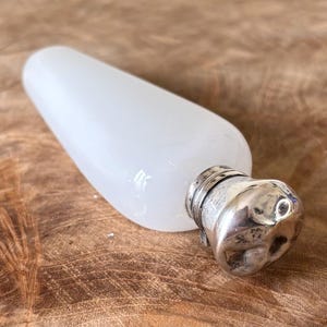 May include: A white glass flask with a rounded body and a silver-coloured metal cap. The flask is lying on a wooden surface. The cap has a textured, organic shape.