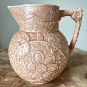 Op de afbeelding: Een perzik-kleurige keramische kan met een bloemenontwerp. De kan heeft een ronde body, een tuit en een handvat met een bloemdetail. Het oppervlak is getextureerd met reliëfbloemen en bladeren. Een decoratief stuk voor de keuken of eetkamer.