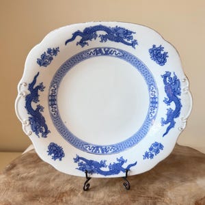 May include: A white ceramic serving bowl with a scalloped edge and blue transferware patterns. The design features four blue dragons, floral motifs, and a geometric border encircling the center. The bowl has integrated handles on the sides.