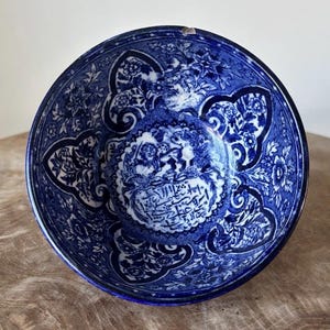 Könnte beinhalten: Eine blau-weiße Keramikschale mit aufwendigen floralen und Rankenmustern. Die Schale hat ein zentrales Medaillon mit Schrift und Figuren. Der Rand hat einen kleinen Chip. Die Schale steht auf einer Holzoberfläche.