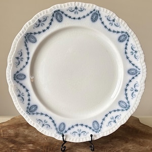 Puede incluir: Plato de cerámica blanca con borde festoneado y borde decorativo azul. El borde presenta medallones ovalados y volutas florales. El plato se exhibe sobre un soporte de metal negro, mostrando su diseño vintage.