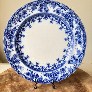 May include: A white ceramic plate with a blue floral pattern. The plate has a scalloped edge and is displayed on a black metal stand. The blue floral design is intricate and covers the rim and inner border.