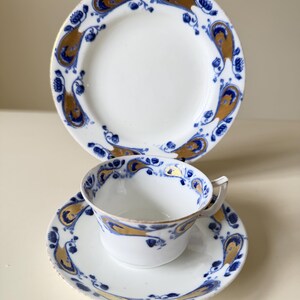 May include: A white porcelain tea set comprising a cup, a saucer, and a small plate. Each piece features a repeating pattern of blue floral motifs and gold-coloured paisley-shaped accents along the edges. The cup has a single handle.