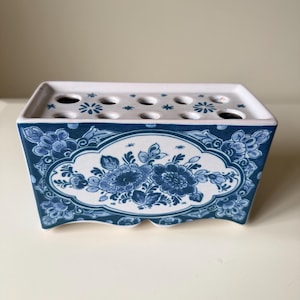 May include: A rectangular ceramic flower frog with a white base and blue floral patterns. The front features an oval medallion containing detailed blue flowers and leaves. The top surface is flat with multiple circular holes for stems.