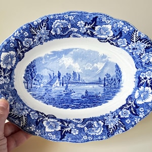 May include: A blue and white oval serving dish with a floral border and a scene of a bridge and houses in the center.