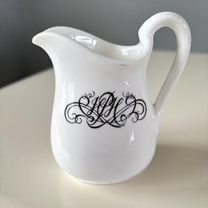 May include: White ceramic creamer with a black cursive monogram. The monogram is a stylized "R" with a flourish.