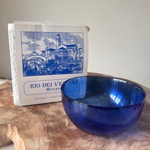 May include: A vibrant blue glass bowl with a curved design. A white paper label with blue illustrations and text is behind the bowl. The label reads "RIO DEI VE MURANO - VENEZIA". The bowl is placed on a wooden surface.