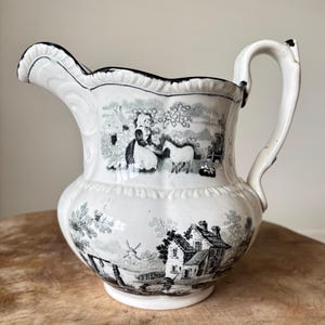 May include: White ceramic pitcher with a black and white pastoral scene. The pitcher features a scalloped rim and a curved handle. The design includes images of people, animals, and buildings, creating a vintage aesthetic.