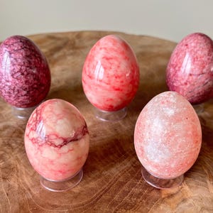 Könnte beinhalten: Fünf dekorative Steineier in Rosa- und Rottönen, jeweils auf einem transparenten Kunststoffständer montiert. Die Eier variieren in Farbe und Maserung, wobei einige einen tiefen Burgunderfarbton und andere ein helleres, marmoriertes Aussehen aufweisen. Sie sind auf einer Holzoberfläche angeordnet.